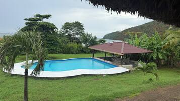 Una piscina con zona verde circundante, un pabellón cubierto y abundante vegetación al fondo.