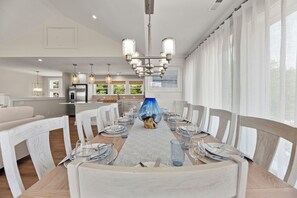 A dining room with a long table set for six, surrounded by white chairs, a chandelier, and a view of the kitchen. - Serendipity: New Build | Saltwater Pool | Hot Tub (Corolla)