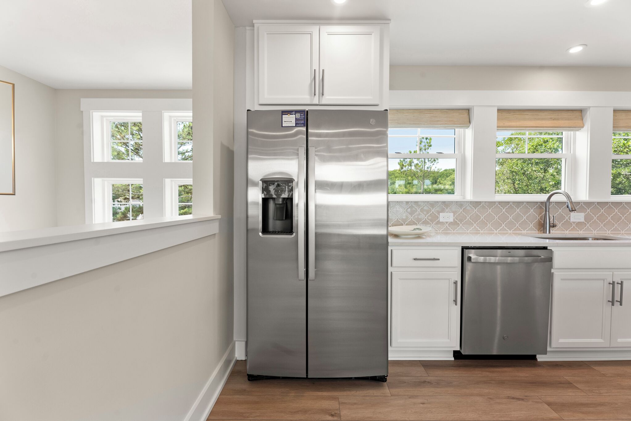 A modern kitchen with white cabinetry, a stainless steel refrigerator, a dishwasher, and a window with a view of greenery.