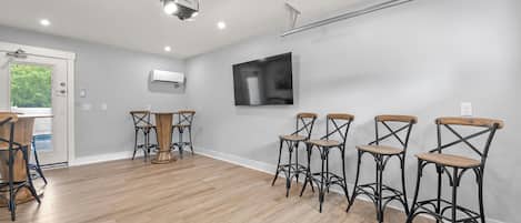 A spacious room with a wooden floor, a round table with chairs, a mounted TV, and an air conditioning unit.