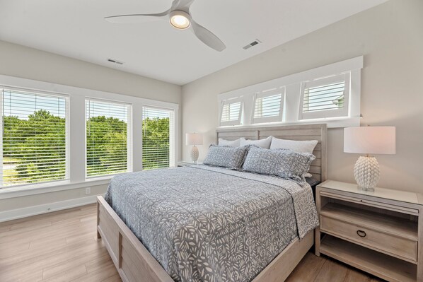 A bedroom with a bed, nightstands, a ceiling fan, and large windows with blinds. - Serendipity: New Build | Saltwater Pool | Hot Tub (Corolla)