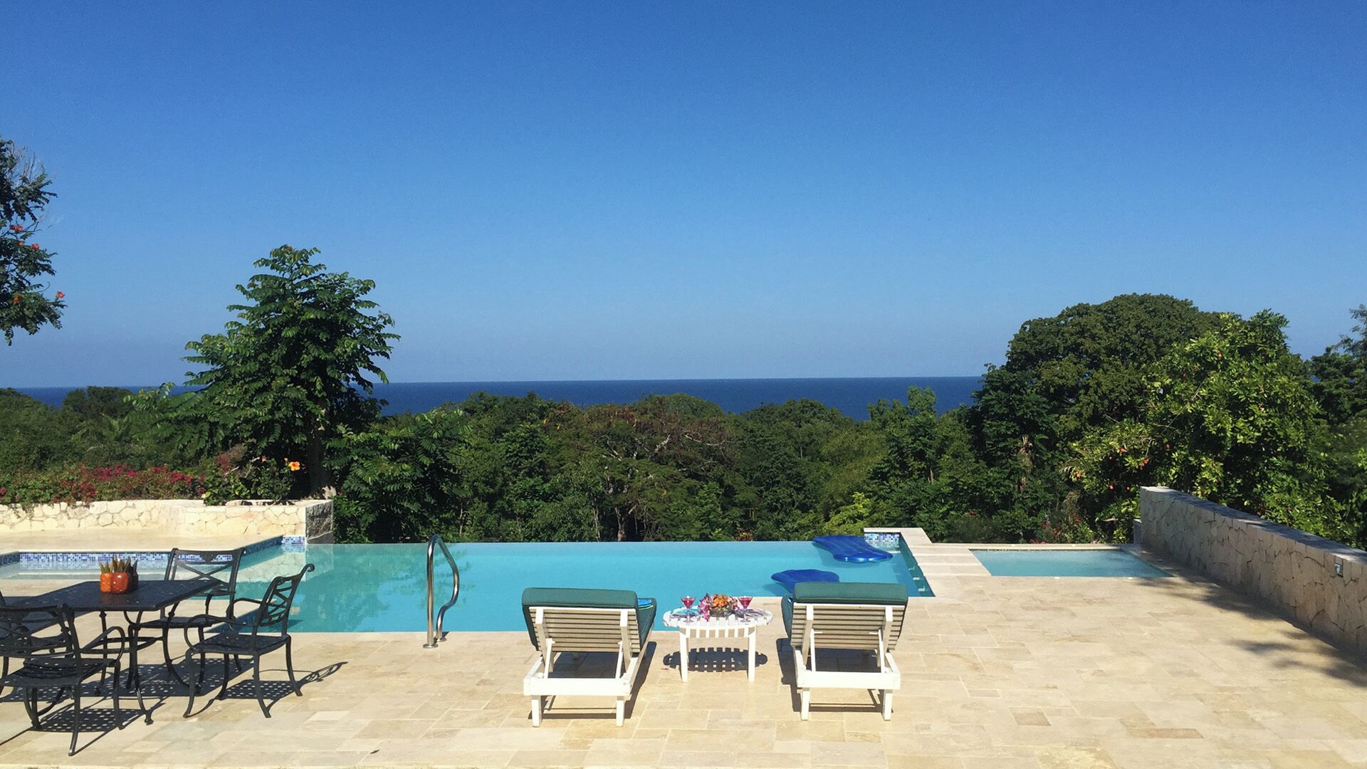 Ein Poolbereich mit Liegestühlen und einem Tisch, umgeben von Bäumen mit Blick auf den Ozean.