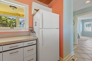 A kitchen with a white refrigerator, a window, and a door leading to another room. - Mr and Mrs Sea: Beach Club Access w/ Pool | Golf Course Views (Nags Head)