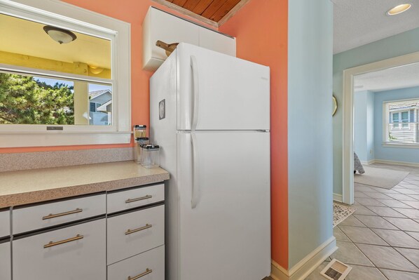 A kitchen with a white refrigerator, a window, and a door leading to another room. - Mr and Mrs Sea: Beach Club Access w/ Pool | Golf Course Views (Nags Head)