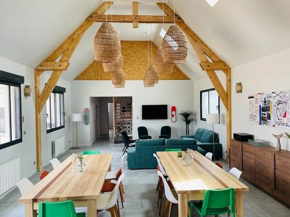 Ein geräumiger Loft mit hohen Decken, Holzbalken, einer Essecke mit einem langen Tisch und einer Wohnbereits mit einer Couch und Sesseln.