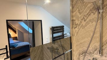 Een moderne badkamer met een douchecabine van glas, een marmeren spatwand, en een zichtbare slaapkamer met dakraam.