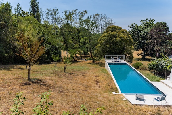 A rectangular swimming pool with a white deck and a metal railing, surrounded by trees and dry grass.
