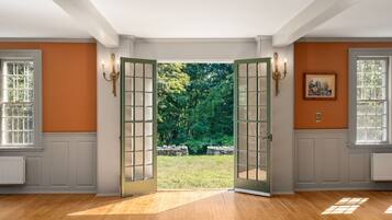 A room with a large wooden door leading to a garden, white walls, and a framed picture on the wall.