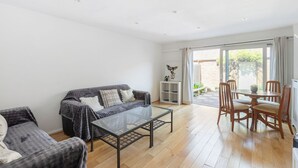 A spacious living room with a sofa, a glass coffee table, and a dining area with chairs and a table. - GuestReady - Charming Retreat with Private Garden (London)