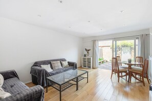 A spacious living room with a sofa, a glass coffee table, and a dining area with chairs and a table. - GuestReady - Charming Retreat with Private Garden (London)