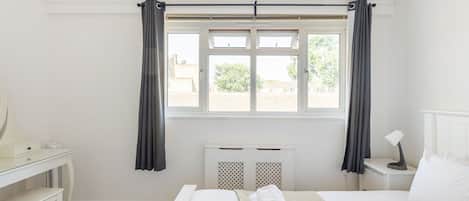 A hotel room with a bed, a desk, a chair, a radiator, and a window with curtains.