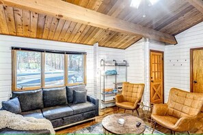 A cozy room with a wooden ceiling, a sofa, two armchairs, a small table, and a window with a view of a road.