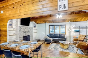 A cozy cabin interior with a dining area, fireplace, sofa, and a sign that says 'HOUSE WE DO FUN'.