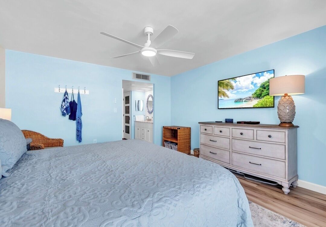 A bedroom with a bed, a ceiling fan, a dresser, a lamp, and a wall-mounted TV.