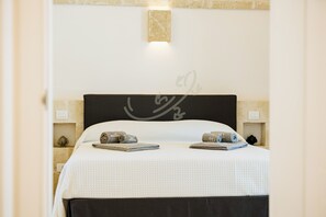 Un lit bien fait, avec une couette blanche et deux serviettes enroulées, une tête de lit sombre au design simple et un petit luminaire fixé au mur au-dessus du lit.