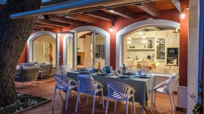 An outdoor dining area with a table set for four, surrounded by chairs, and a view into a modern kitchen through glass doors. - Villa Fiola 8, Emma Villas (Ancona)