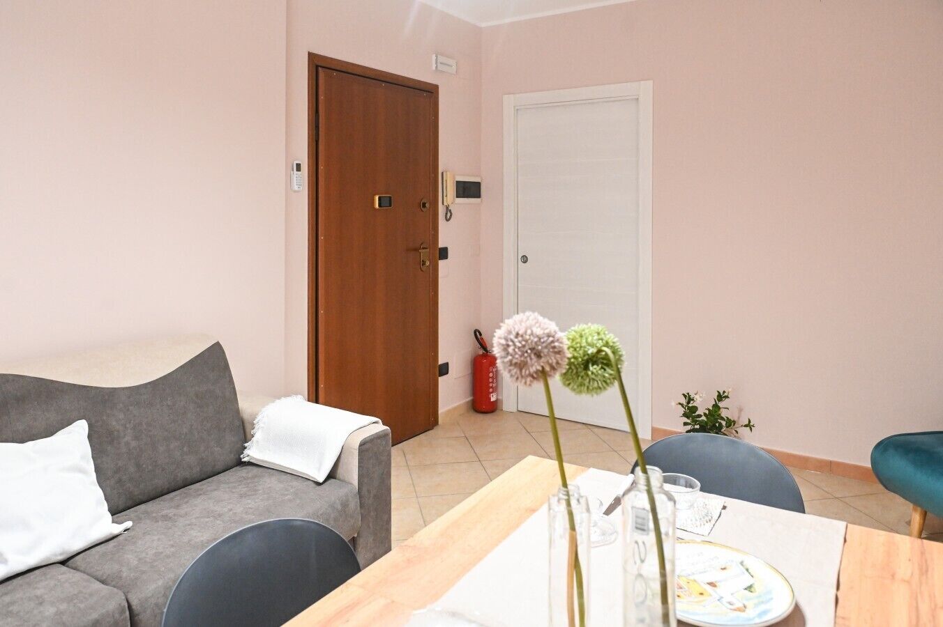 A room with a grey sofa, a wooden door, a white door, and a small table with a vase and a plate.