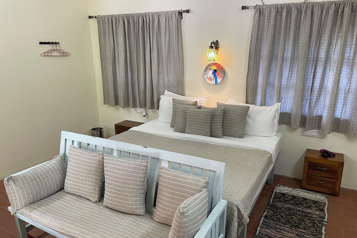 A bedroom with a bed, a bedside table, a nightstand, a wall-mounted light, a hanging light, a wall-mounted outlet, a coat rack, and a window with curtains.