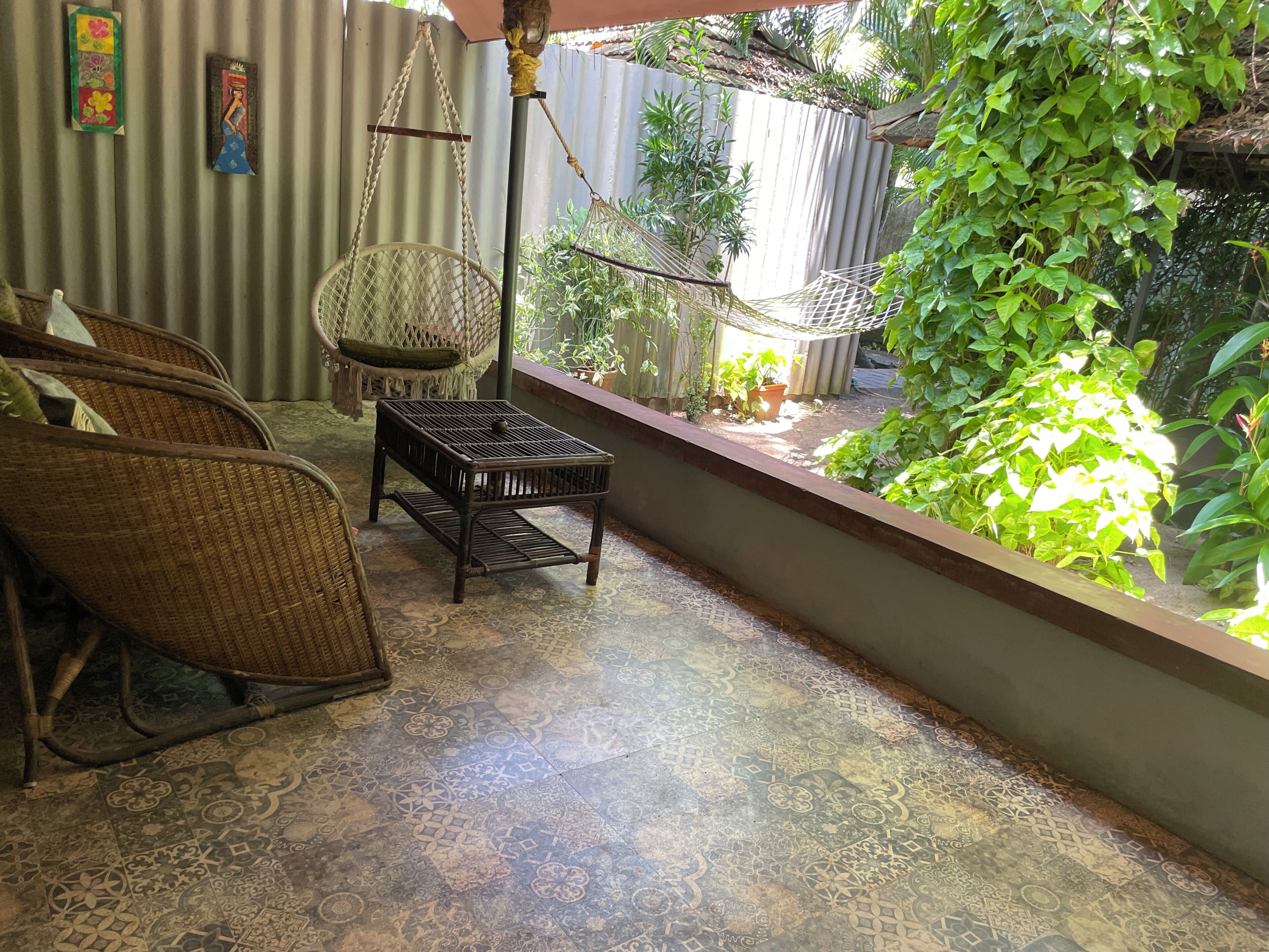 Habitación con muebles de mimbre, piso estampado y vista a un jardín con plantas y una hamaca.