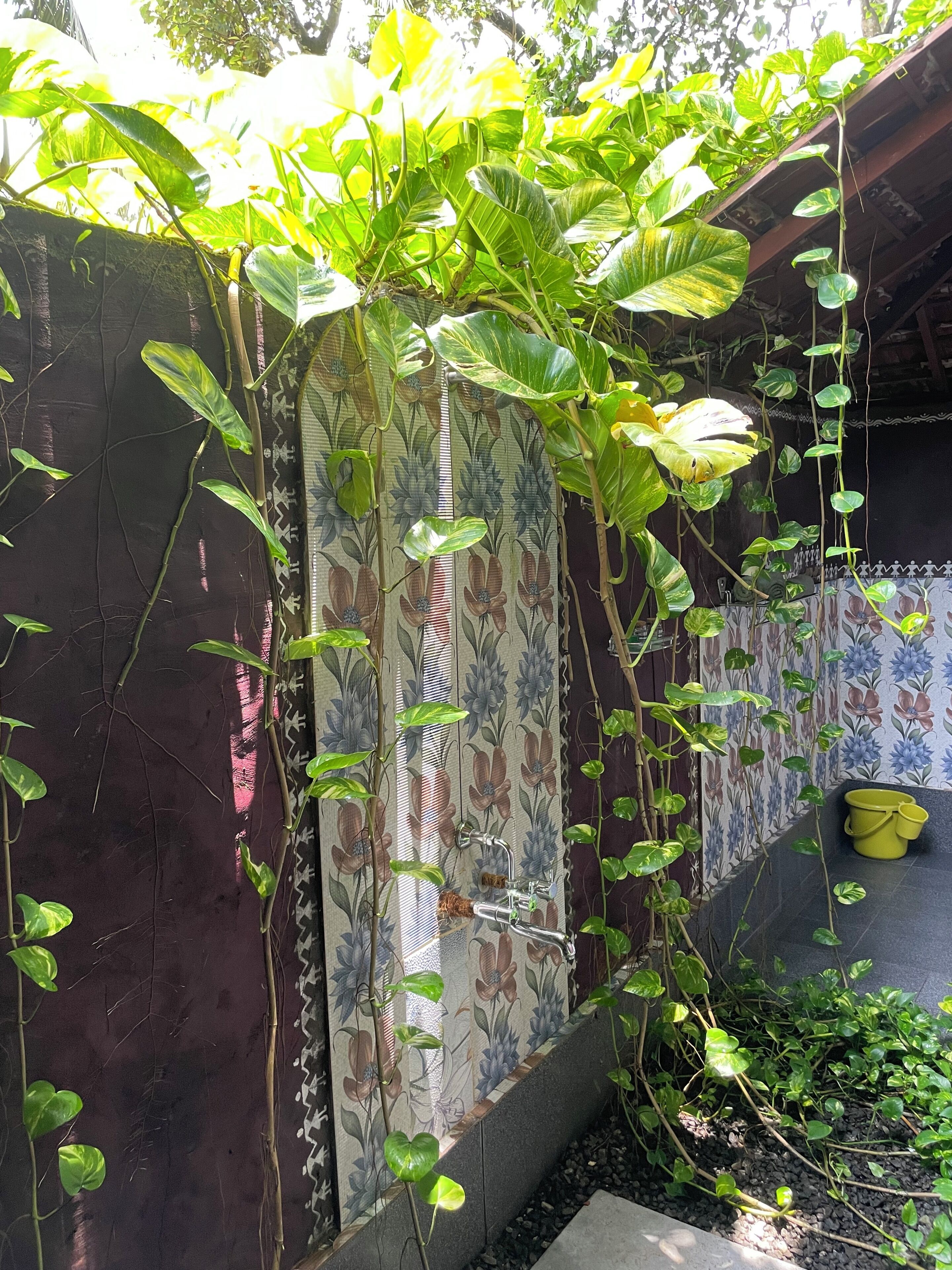 Een tuin met klimplanten, een betegelde muur met bloemenmotieven en een gele emmer.