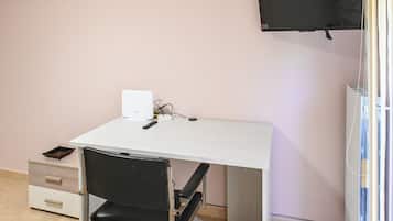 Una habitación pequeña con un escritorio blanco, una silla negra, un televisor montado en la pared y un radiador.