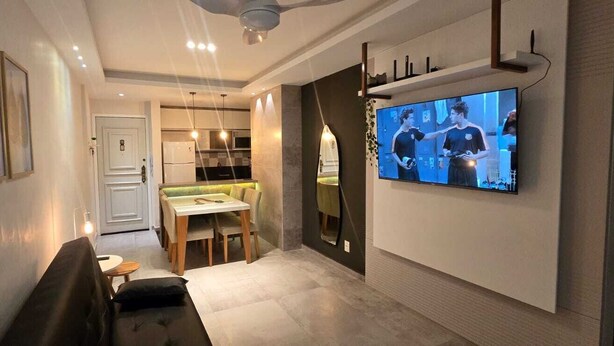 A modern living room with a flat-screen TV mounted on the wall, a dining area with a table and chairs, and a sofa. - Decorated corner near the beach in Jardim Camburi (Vitória)