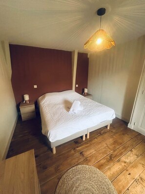 A bedroom with a wooden floor, a bed with a white bedspread, a bedside table with a lamp, and a woven basket. - Les 3 Tilleuls* Charme & Jardin in the heart of Sarlat (Sarlat-la-Canéda)