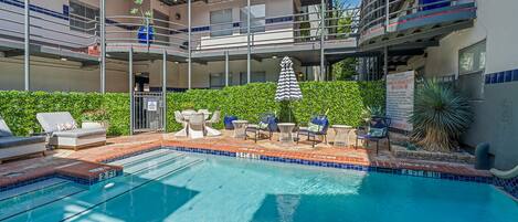 A swimming pool with lounge chairs and umbrellas, surrounded by a multi-story building with balconies.