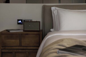 Una cama con ropa de cama blanca, una mesita de noche de madera con un altavoz y un libro, y un reloj digital que muestra la hora.