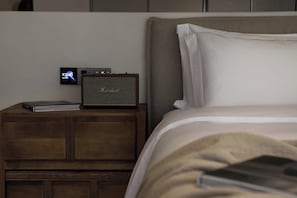 A bed with white bedding, a wooden nightstand with a speaker and a book, and a digital clock displaying the time.