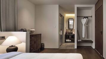 A hotel room with a bed, a bedside lamp, a wooden dresser, a bathroom with a mirror, and a robe hanging on a rack.