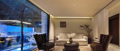 A modern living room with a large window, a white sofa, two leather armchairs, a coffee table, and a vase with plants.