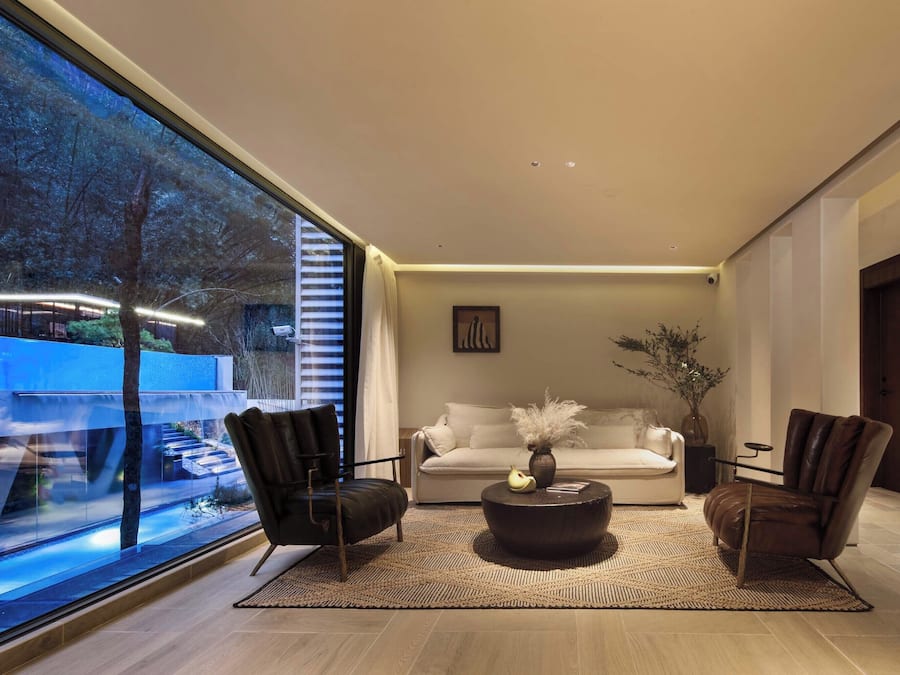 A modern living room with a large window, a white sofa, two leather armchairs, a coffee table, and a vase with plants.
