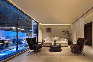 Un salón moderno con un ventanal grande, un sofá blanco, dos sillones de cuero, una mesa de centro y un florero con plantas.