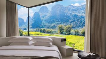 A bedroom with a large window, a bed, a bedside table, and a view of a mountainous landscape.