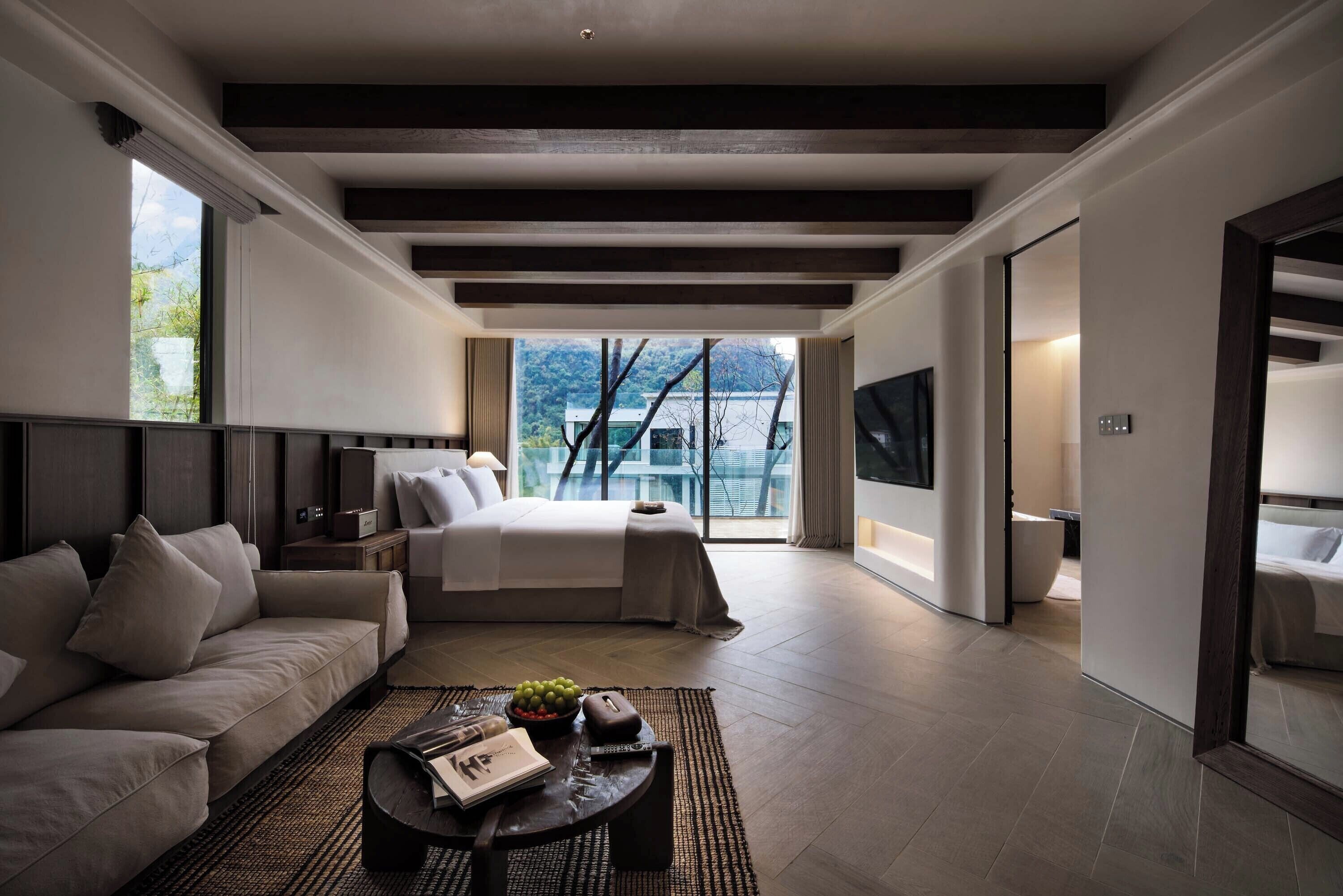 A modern hotel room with a large bed, a sofa, a coffee table with a magazine, and a view of the outdoors.