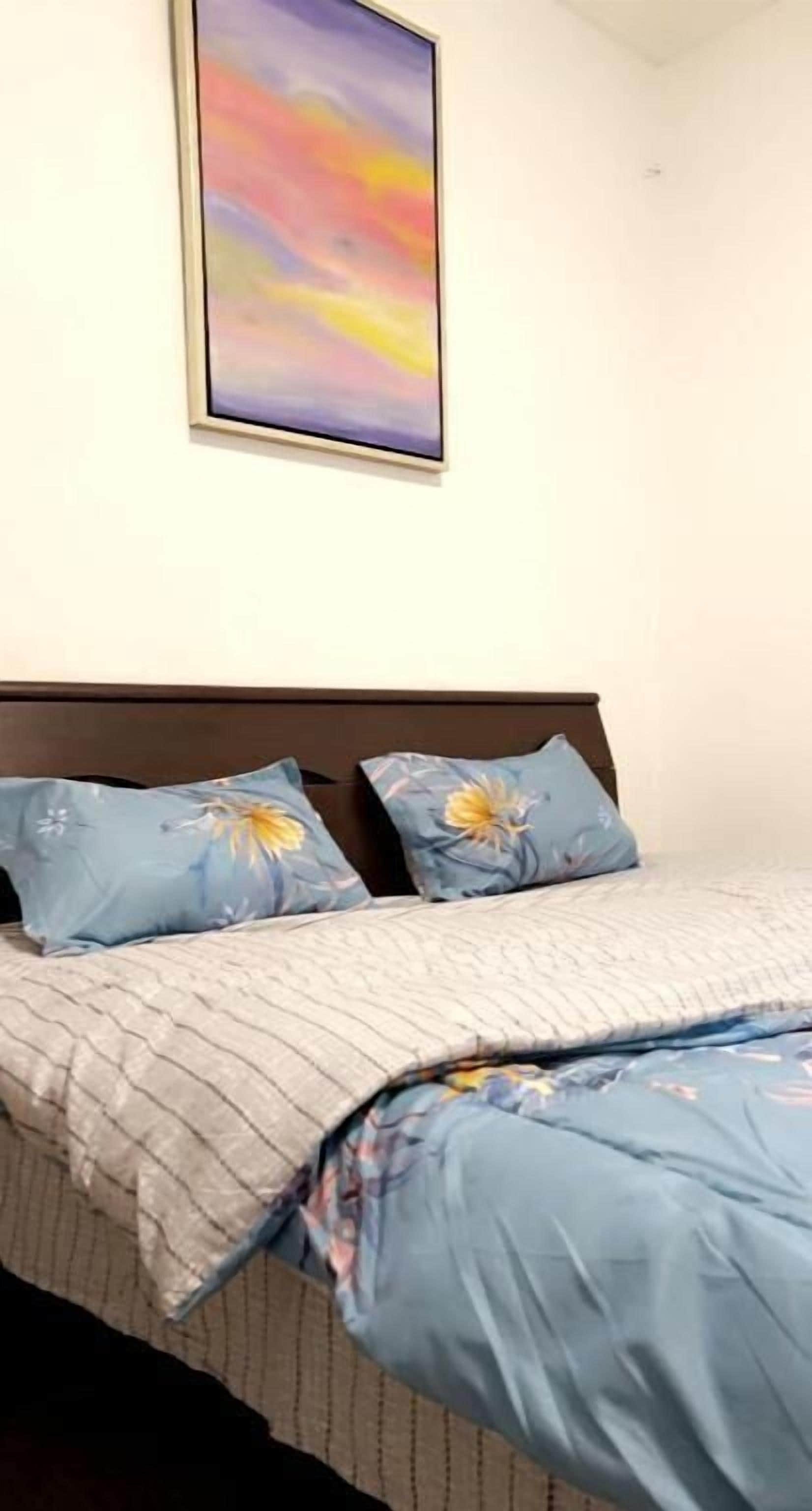 A bed with a floral bedspread and matching pillows, a framed picture on the wall, and a plain white ceiling.
