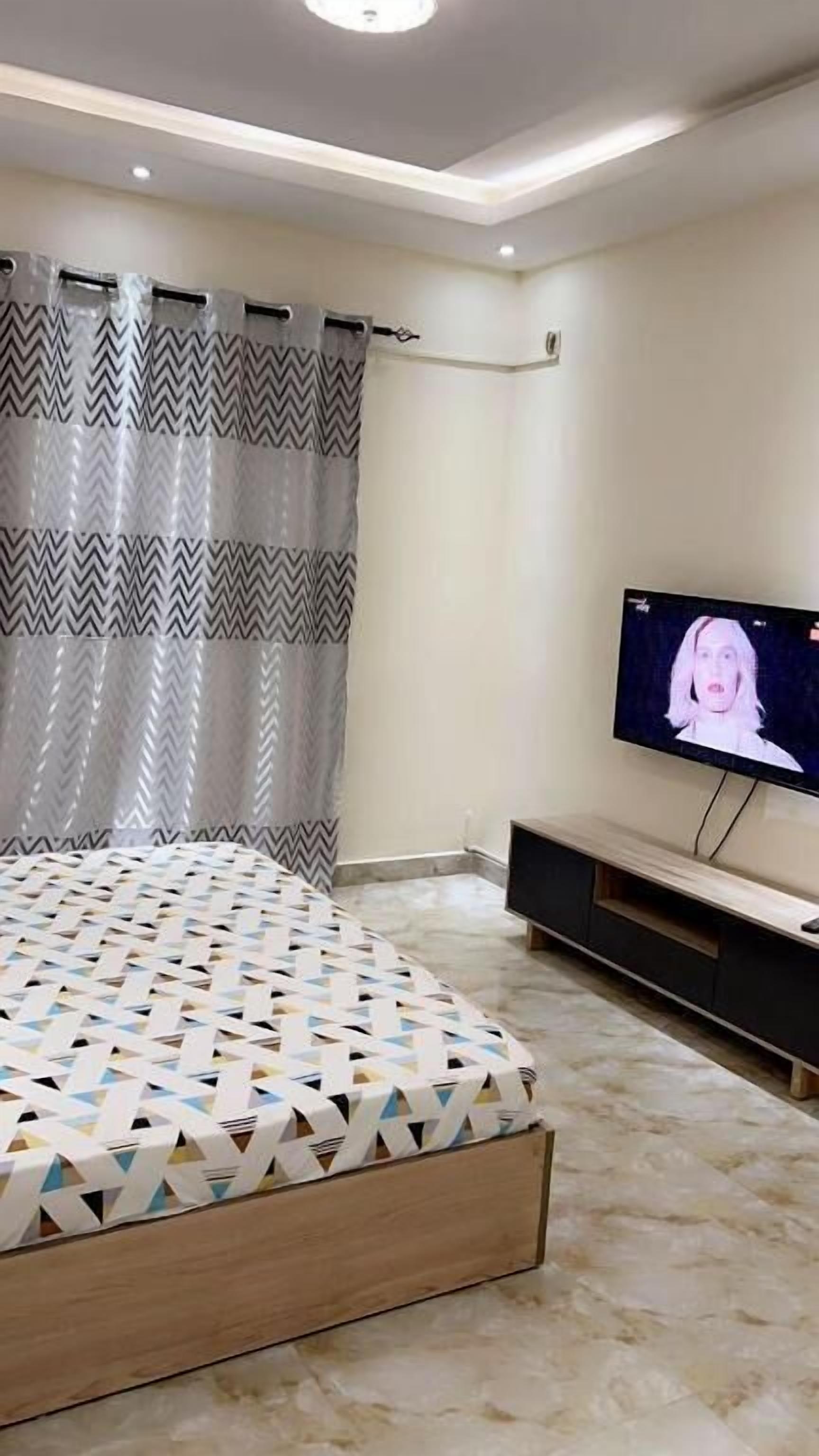 Un dormitorio con cama, televisor, cortina y estante fijado a la pared.