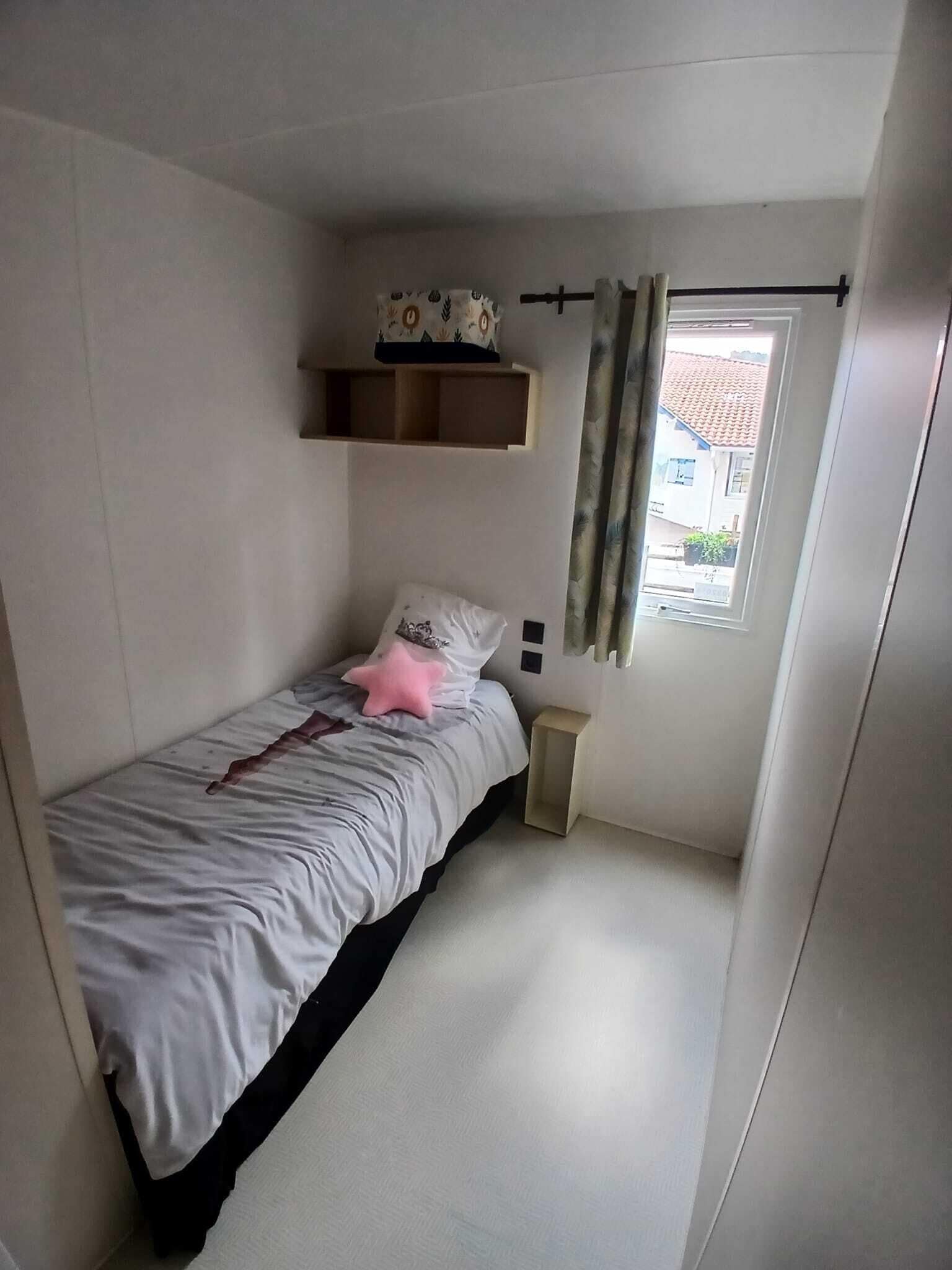 A small, single-bed room with a window, a bedside table, and a shelf above the bed.