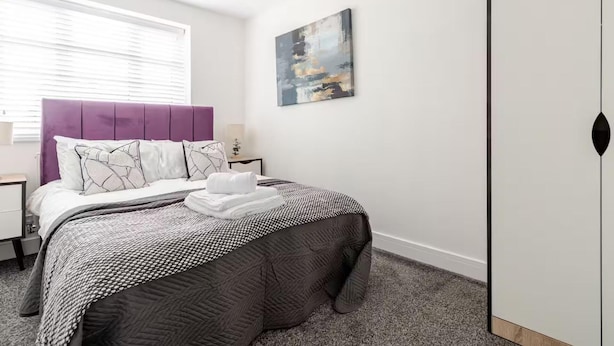 A bedroom with a bed, a nightstand, a bedside table, a window with blinds, a painting on the wall, and a wardrobe. - Luxurious Stockport Retreat - 4 Bedrooms (Stockport)