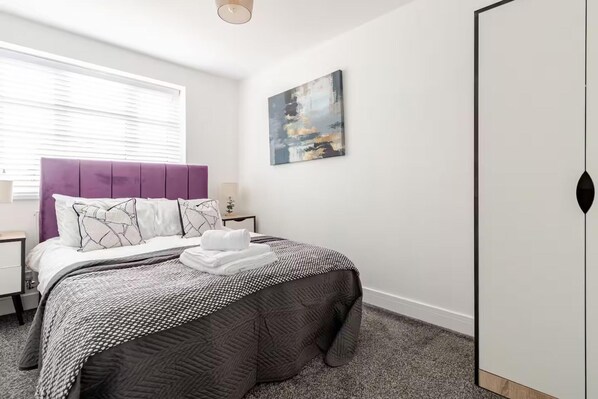 Um quarto com cama, mesa de cabeceira, armário e uma janela com persianas, além de um quadro na parede.