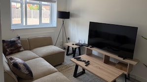 A living room with a sectional sofa, a TV on a stand, a floor lamp, and a window with blinds. - Luxurious Stockport Retreat - 4 Bedrooms (Stockport)