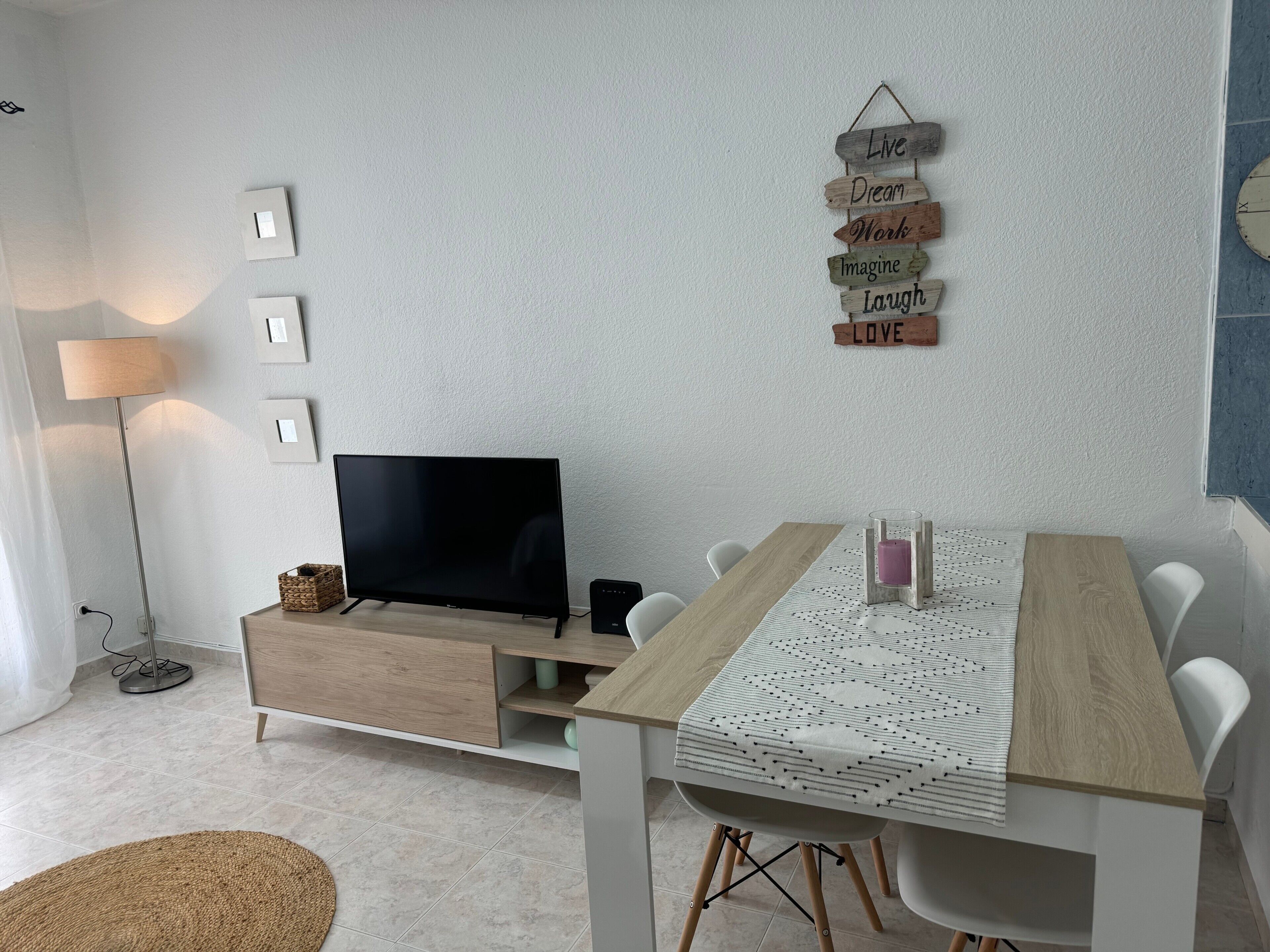 A modern living room with a flat-screen TV, a wooden dining table, and a wall-mounted sign with motivational phrases.