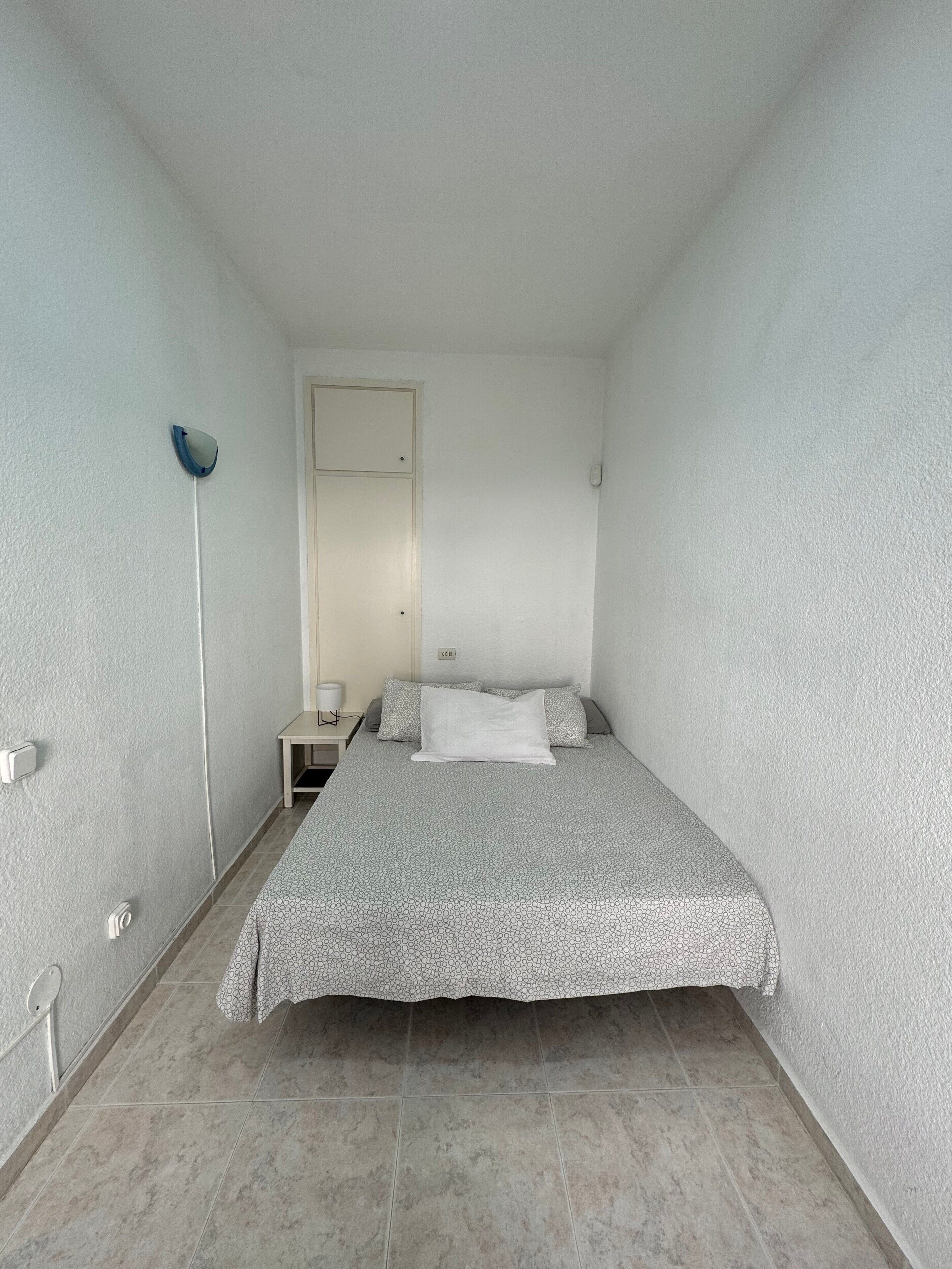 A small, single-bed room with a plain white wall, a bedside table with a lamp, and a door.