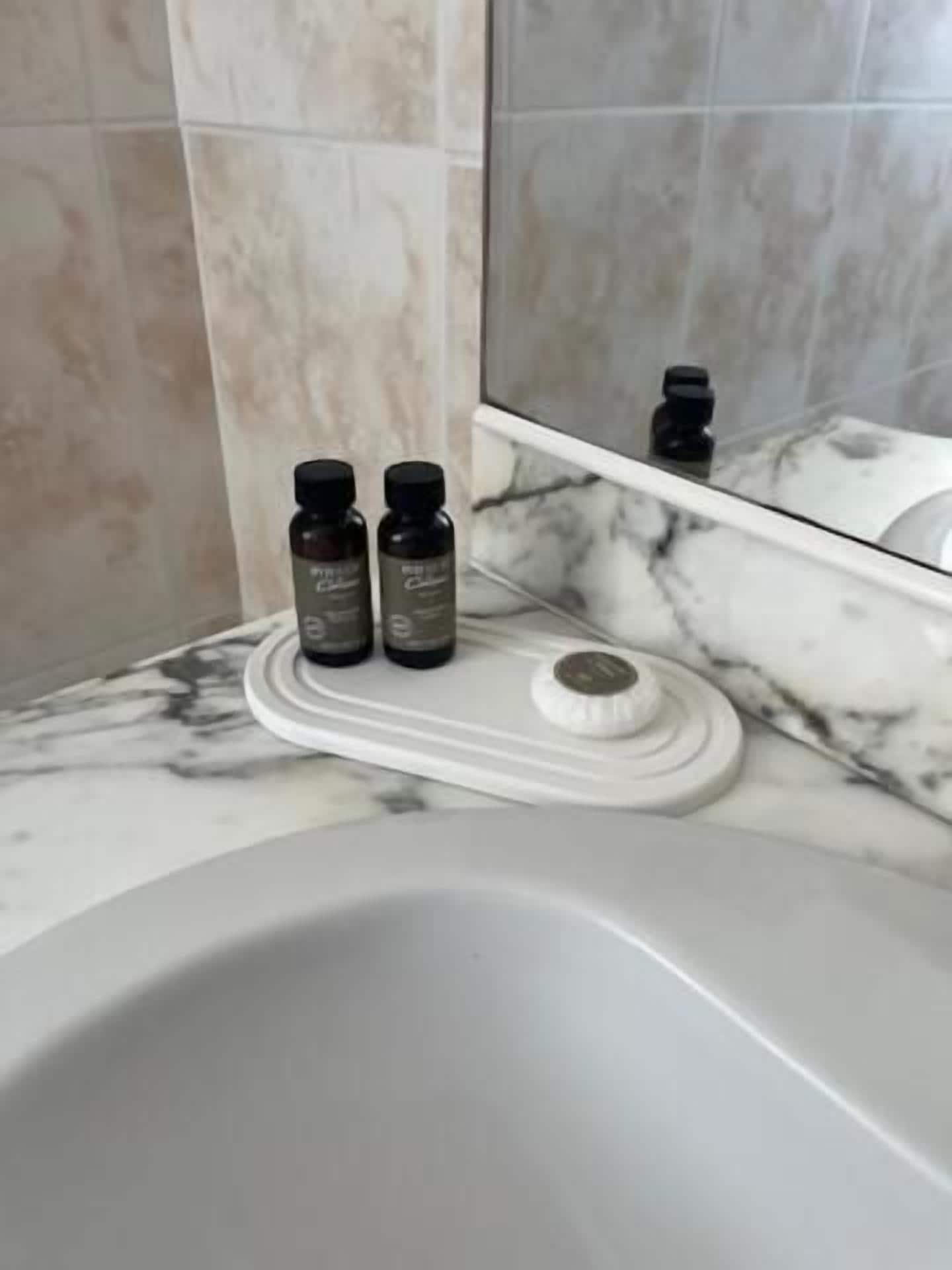 Baño con encimera de mármol, lavamanos blanco y dos botellas pequeñas sobre una bandeja.