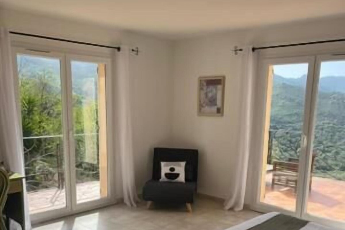 A bedroom with a bed, a small table, and a chair. There is a balcony with a view of the outdoors.