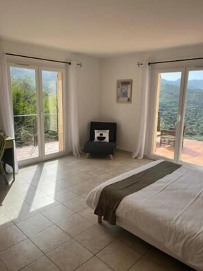 A bedroom with a bed, a small table, and a chair. There is a balcony with a view of the outdoors.
