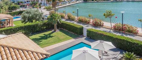 A swimming pool with a wooden deck, surrounded by neatly trimmed hedges and palm trees.