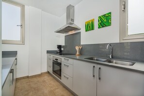Een moderne keuken met een fornuis, oven, spoelbak en een afzuigkap. Er zijn kastjes met handgrepen, een raam en een deur.