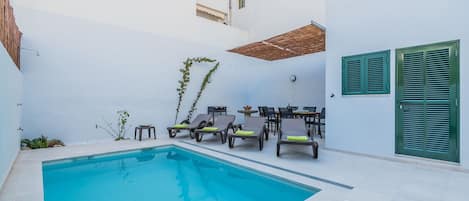A swimming pool with lounge chairs and a table, surrounded by white walls and a tiled floor.